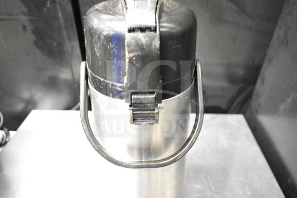 Stainless steel air pot with black handle and push-button lid, showing signs of wear. Ideal for storing and dispensing beverages.
