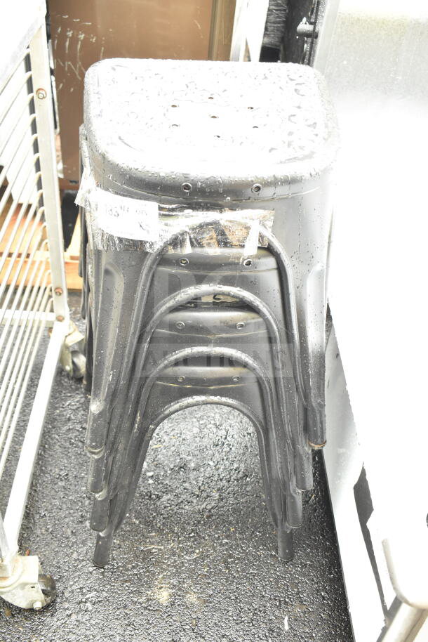 Four brand new gray Tolix style stools with minor scratches and dents, stacked, wet surface, bagged label visible.