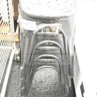 Four brand new gray Tolix style stools with minor scratches and dents, stacked, wet surface, bagged label visible.