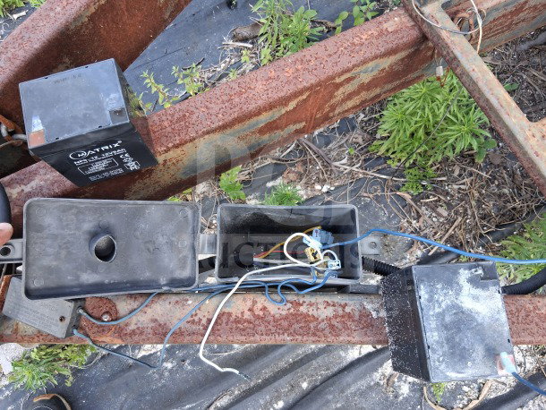Rusty metal framework with exposed wiring and electrical components, including a Matrix brand box. Outdoor setting with greenery.