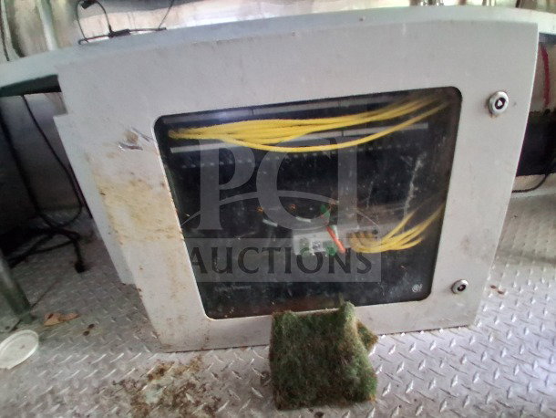 The image shows a white metal panel with a dirty glass window, exposing yellow cables inside. The panel is mounted on a textured metal floor.