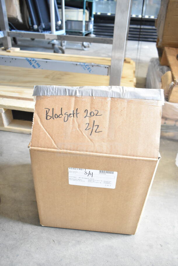 Cardboard box with handwritten "Blodgett 202 2/2" text and a label indicating shipping information, on a warehouse floor.