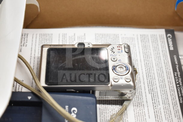 Canon digital camera, silver body, backside view with screen and control buttons. Includes neck strap and Canon documents.