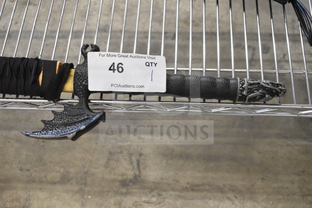 Samurai sword in sheath with dragon-themed hilt, black wrapping, and auction tag marked "46", placed on metallic shelf.
