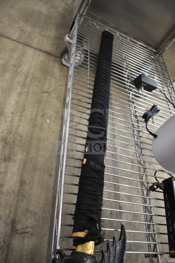 Samurai sword in black sheath, visible wear on handle, ornate guard, displayed on metal cart.