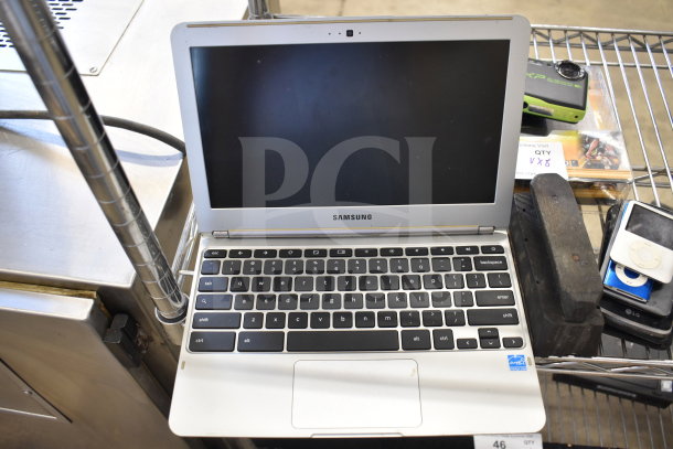 Samsung laptop with black keyboard and screen, accompanied by four smart devices, on metal cart. Items in used condition.