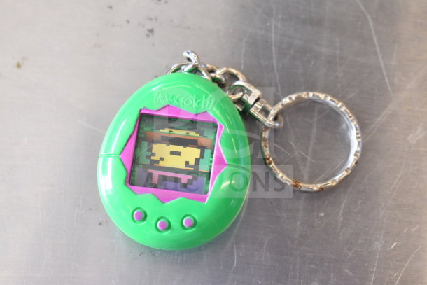 Green Tamagotchi with keychain, featuring pixelated character on screen. Slight wear visible on metal ring.