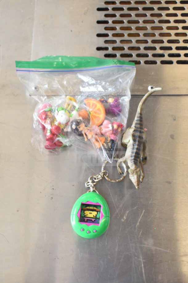A collection of 15 items including a green Tamagotchi, dinosaur figure, various figurines in a bag, and hand sanitizer cozies.