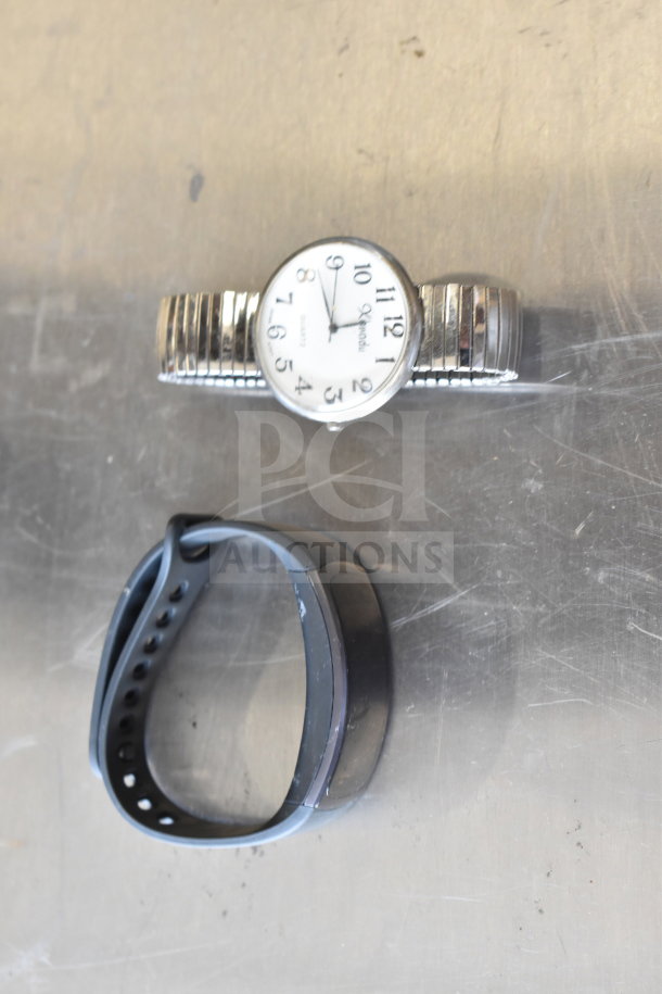 Two watches on a metal surface; a silver Xpedi quartz with a white face, and a black fitness band. Moderate wear visible.