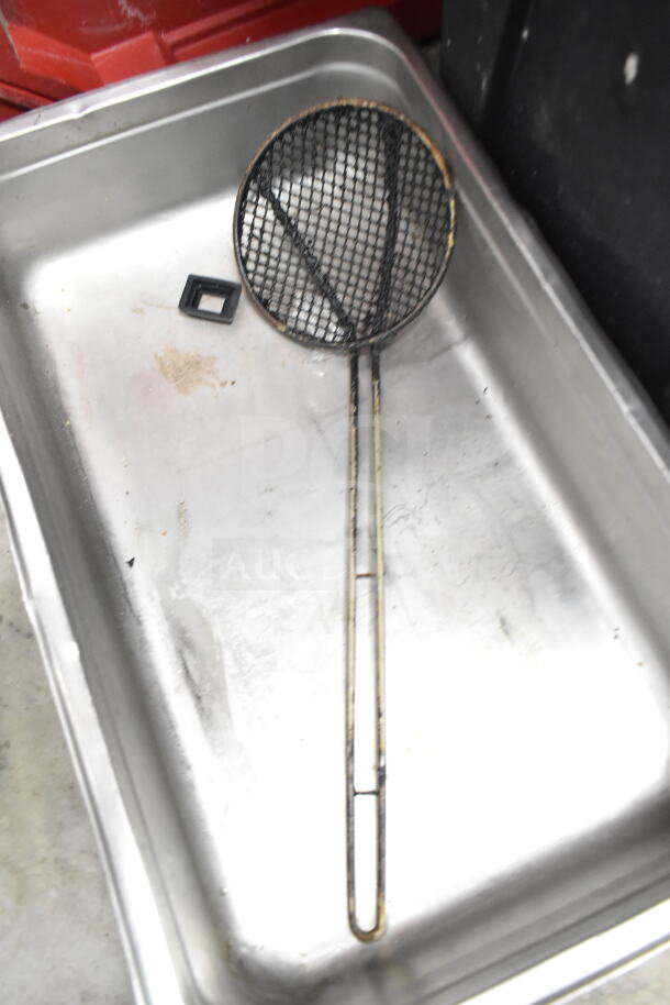 Metal serving basket with long handle, placed in a stainless steel container; shows signs of wear.