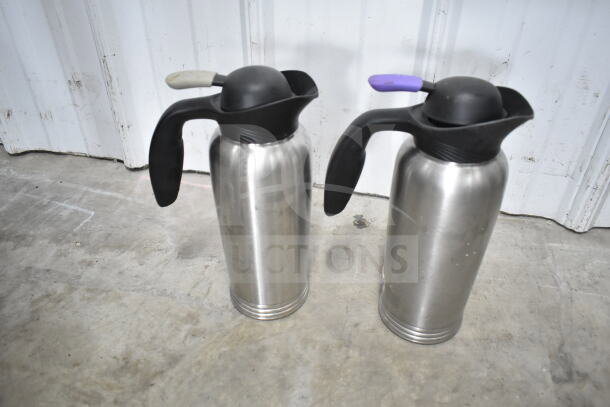 Two stainless steel air pots with black handles, one with a purple grip and the other with a grey grip, on a concrete floor.