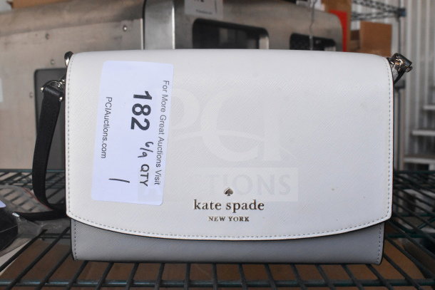 Kate Spade New York white purse with black strap, branded logo on front, attached auction tag, and metallic accents.