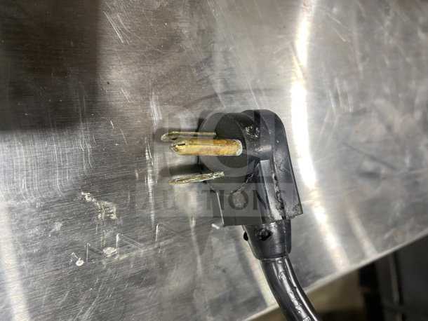 A black electrical cord plug with three prongs is shown against a metallic surface, highlighting its condition.