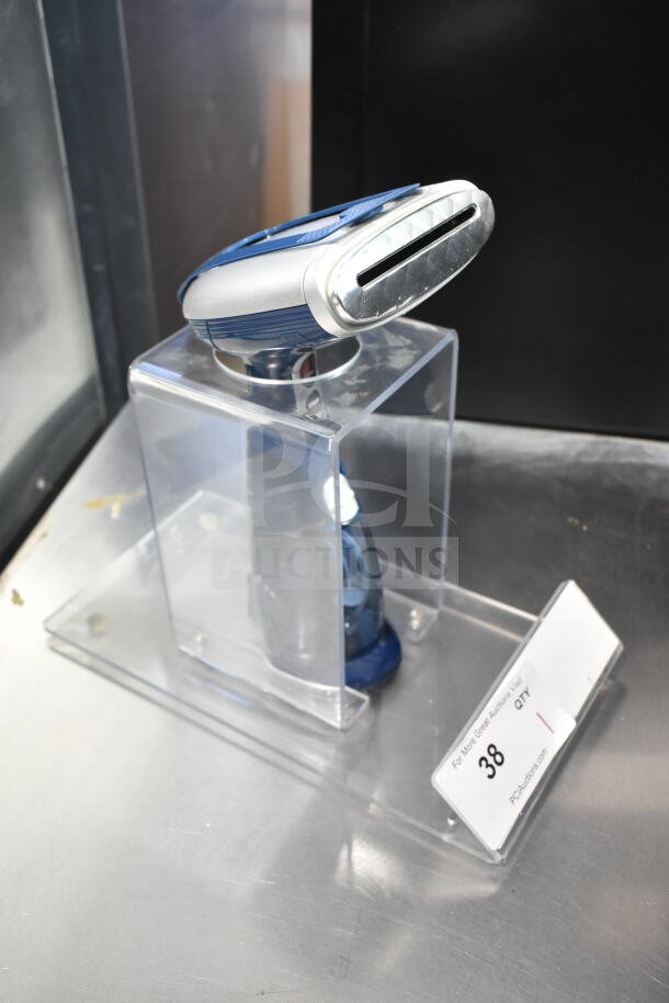 Compact handheld steamer in transparent stand, features a chrome nozzle and blue grip. Auction label indicates "Lot 38."