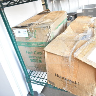 Two boxes on a shelf, labeled "EcoChoice 8 oz. Kraft Coffee Cup Sleeve" and "Huhtamaki 10-1/4” Plastic Plates." Boxes appear taped and in used condition.