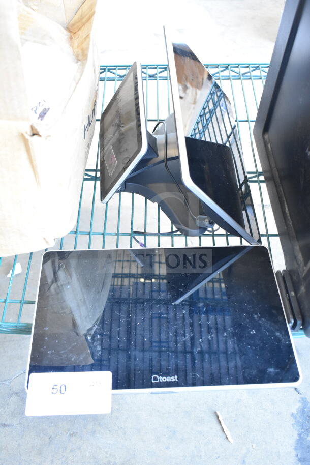 Two Toast POS monitors on a metal rack, showing black screens. Condition appears used, with small visible marks.