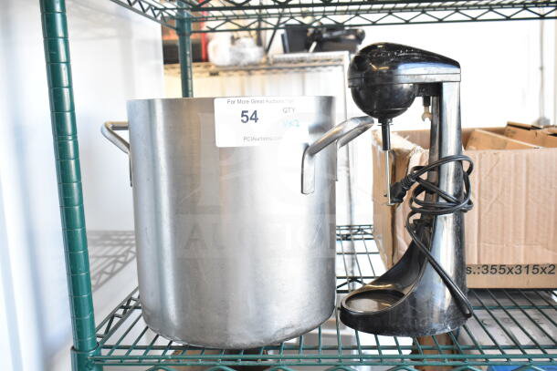 Alt text: Large stainless steel stock pot with handles and ST789 black milkshake mixer on metal shelf, auction label visible.