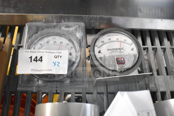 Two Magnehelic gauges, one in packaging. Measures pressure in pascals/inches of water. Auction tag reads "144 QTY 2X".