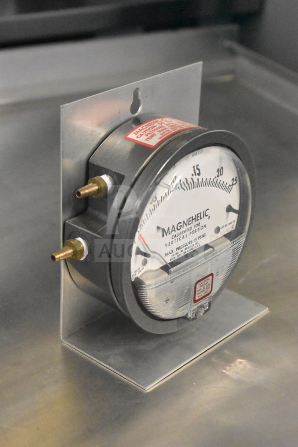 Magnehelic gauge displaying pressure calibration, mounted on metal bracket. Features include dual brass fittings, clear dial.