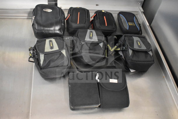 Nine camera bags with two cameras, including Nikon Coolpix and Garmin Nuvi. Black bags, mixed materials, some branded Targus.