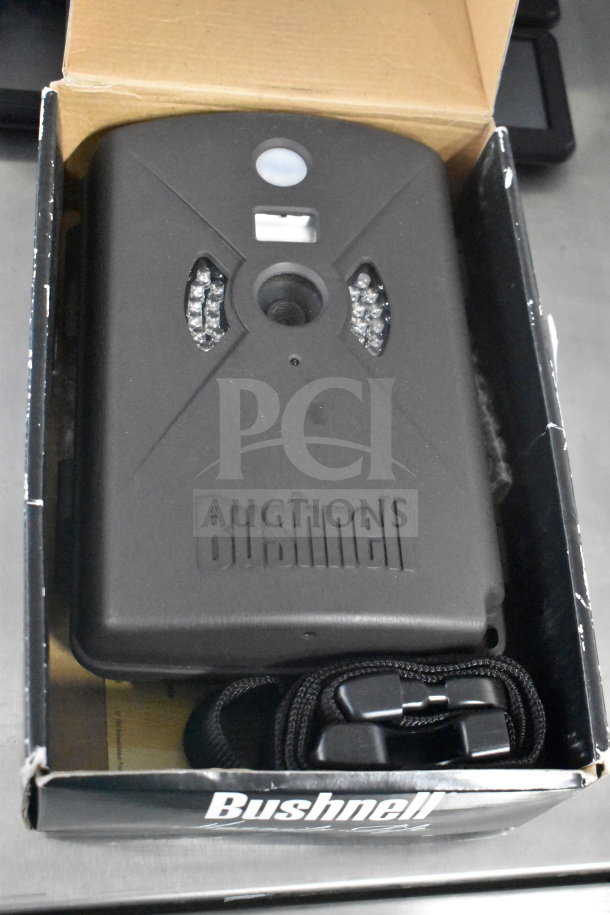 Bushnell trail camera in box, new condition, includes strap; part of auction lot with 4 Garmin GPS devices.