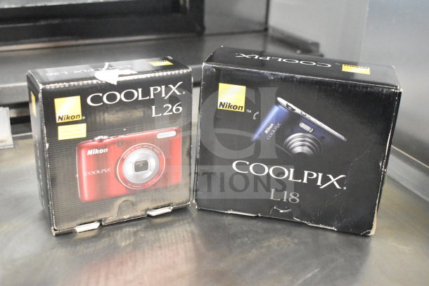 Nikon Coolpix L18 and L26 digital cameras in original boxes. Boxes show camera images and branding.