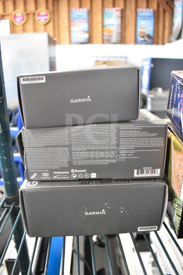 Three Garmin GPS devices in original boxes, showcasing brand logos and features like Bluetooth and Foursquare integration. Mint condition.