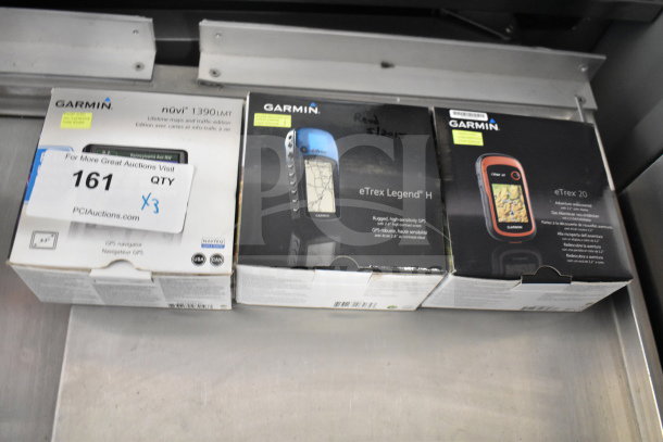 Three Garmin GPS devices in original boxes: eTrex Legend H, eTrex 20, Nuvi 1390LMT. Auction tag indicates triple bid value.
