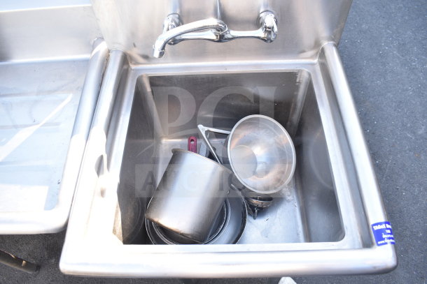 Commercial single bay sink in stainless steel, with faucet, showing light use, contains metal kitchenware; label on side.