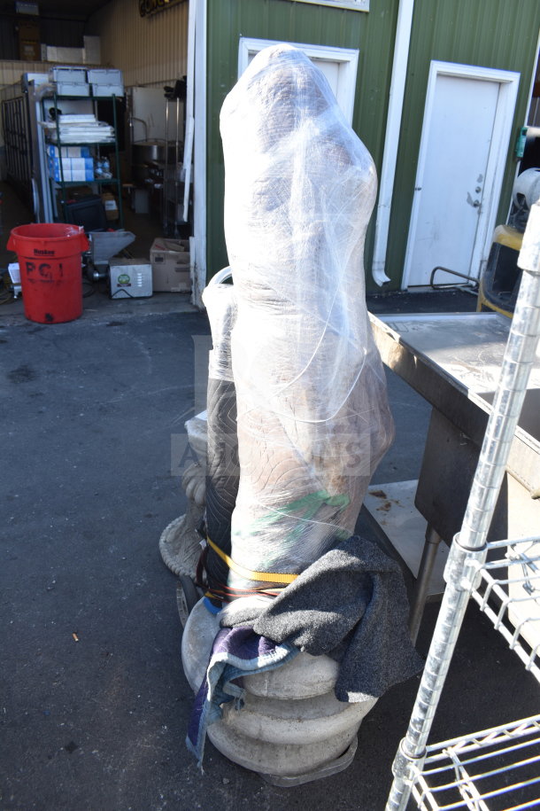 Wrapped statue on a pedestal, covered in protective plastic and padding, displayed outdoors for auction.