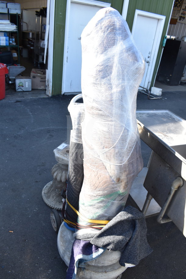 Wrapped statue on base, covered in protective materials and shrink wrap, outdoors near storage area. Condition unknown.