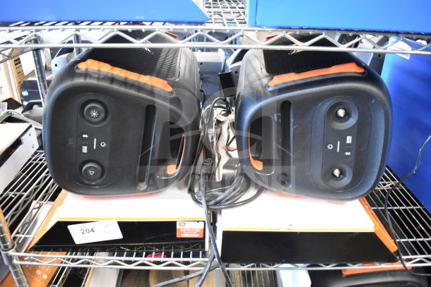Two JBL PartyBox portable Bluetooth speakers, black with orange accents, on a shelf. Cords and labels visible.