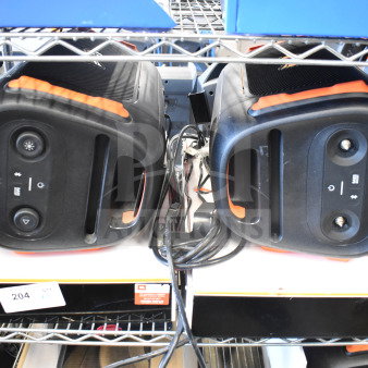 Two JBL PartyBox portable Bluetooth speakers, black with orange accents, on a shelf. Cords and labels visible.