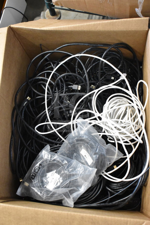 Lot of cables in a cardboard box, with mixed black and white wires. Includes cables in plastic packaging, some labeled.