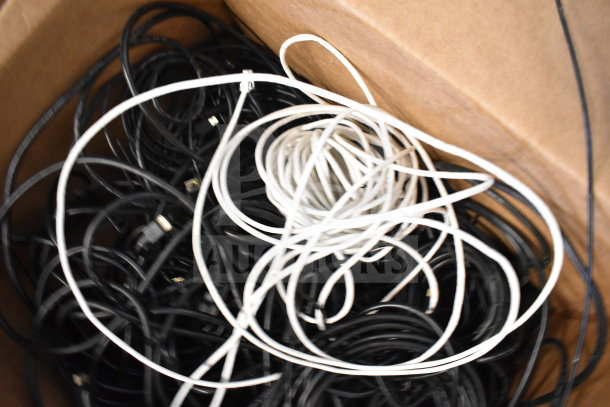 Lot of mixed cables in a cardboard box, featuring both black and white wires, suitable for various electronic uses.