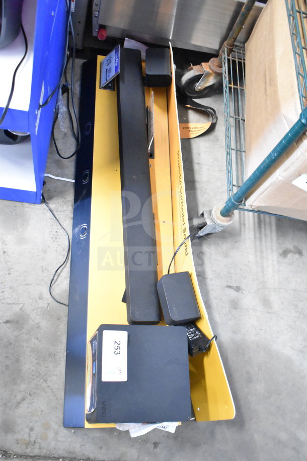 Vizio sound bar and speaker on yellow store display, cords attached. Tags indicate item number 253.