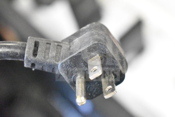 Close-up of a black electrical plug with visible prongs and markings. Part of store display items with routers and headphones.