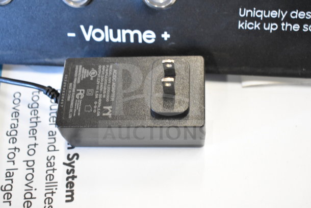 Black AC/DC adapter with visible specifications label, part of store display setup featuring routers, volume signage.