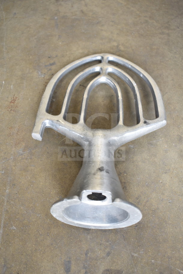 Metal paddle attachment for mixer on concrete surface, showing signs of wear, ideal for kitchen mixing needs.