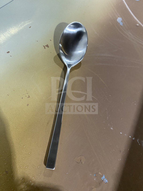 Brand new commercial stainless steel teaspoon with a shiny finish, shown on a metallic surface.