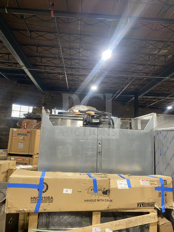 A large industrial refrigerator in a warehouse setting. Visible model number HNC-210BA on a cardboard box in front, suggesting it contains parts or accessories. The refrigerator appears in new or good condition, wrapped and stored with care labels visible.