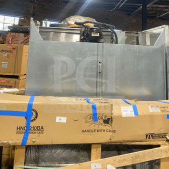A large industrial refrigerator in a warehouse setting. Visible model number HNC-210BA on a cardboard box in front, suggesting it contains parts or accessories. The refrigerator appears in new or good condition, wrapped and stored with care labels visible.