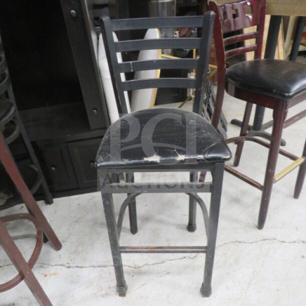 Bar height black metal chair with black cushioned seat, showing wear. Ladder back design. Ideal for bars or kitchens.