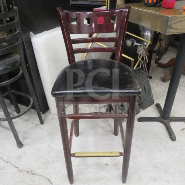 Wooden bar height chair with black cushioned seat, ladder-style back, and footrest. Good condition. Model: 2XBID.