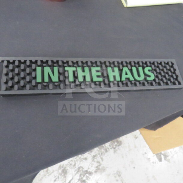 Black rubber bar mat with raised text "IN THE HAUS" in green. Rectangular with textured surface. Good condition.