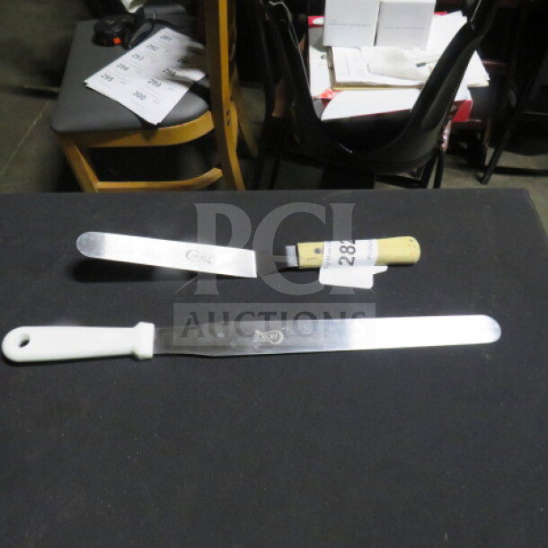 Assorted commercial spreaders on table, metal blades, white plastic and wooden handles, item tags visible.