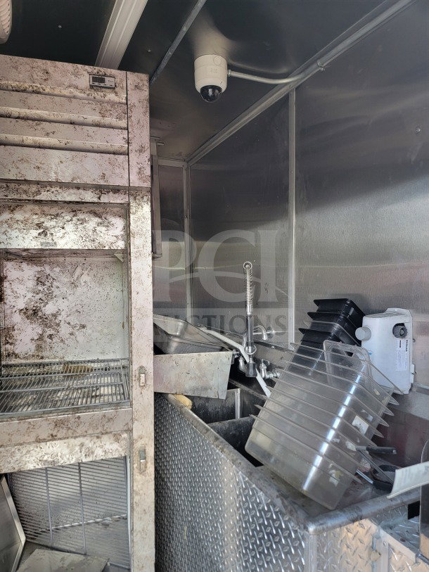 Interior of a used 2021 Rock Solid Cargo 8.5' x 24' mobile kitchen trailer. Features stainless steel shelving, a sink, and a security camera. Visible wear and grime present.