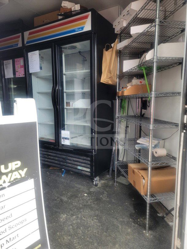 Dual glass-door True refrigerator inside SDG 8.5' x 20' trailer, used for retail merchandising. Metal shelving visible on right.