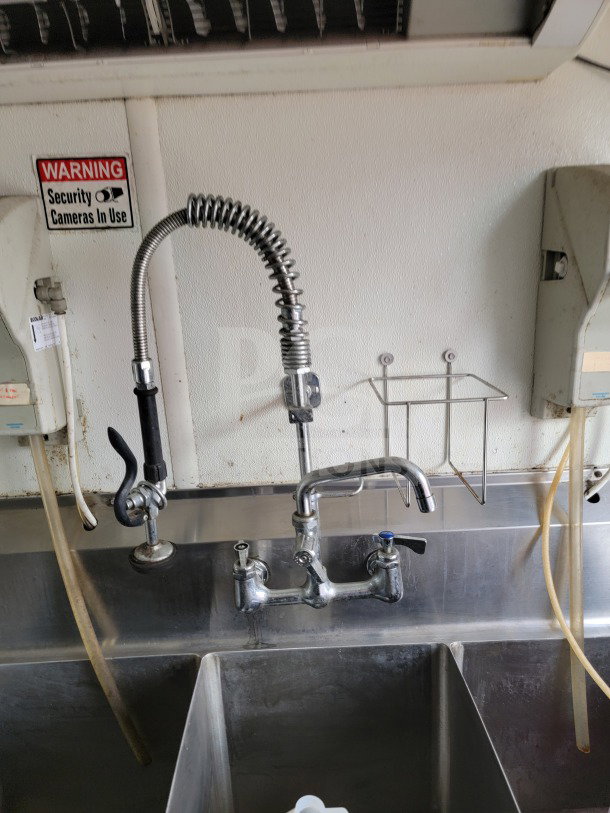 Stainless steel sink area in Diamond Cargo mobile kitchen trailer with high-arc faucet and "Security Cameras In Use" sign.
