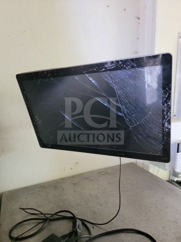 Cracked computer monitor on a stand with visible power cable on a speckled surface.